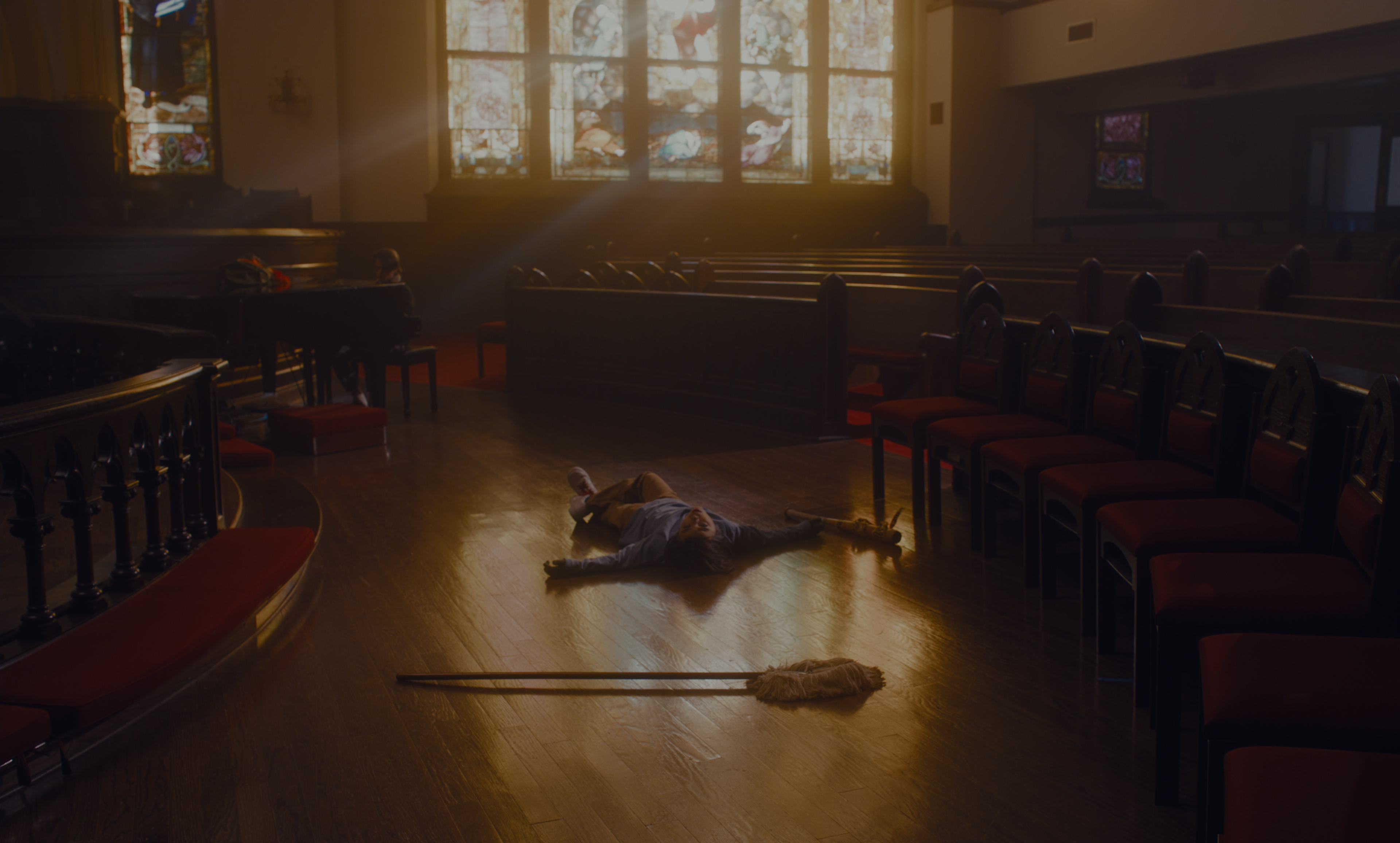 A person lying on the wooden floor of a church aisle beside a mop, warm light through the stained glass