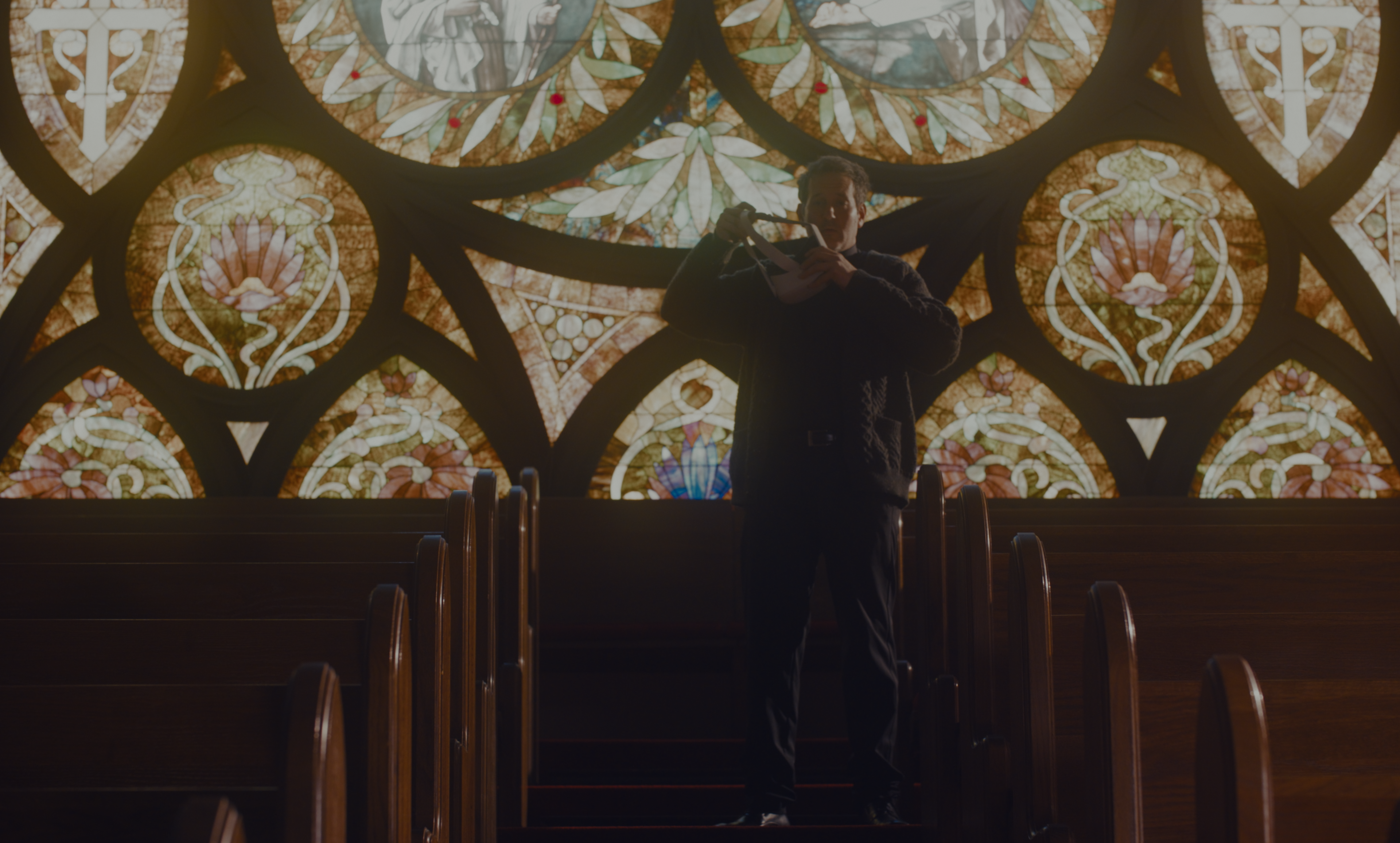 A man adjusting his collar in the church pews beneath a large stained-glass window