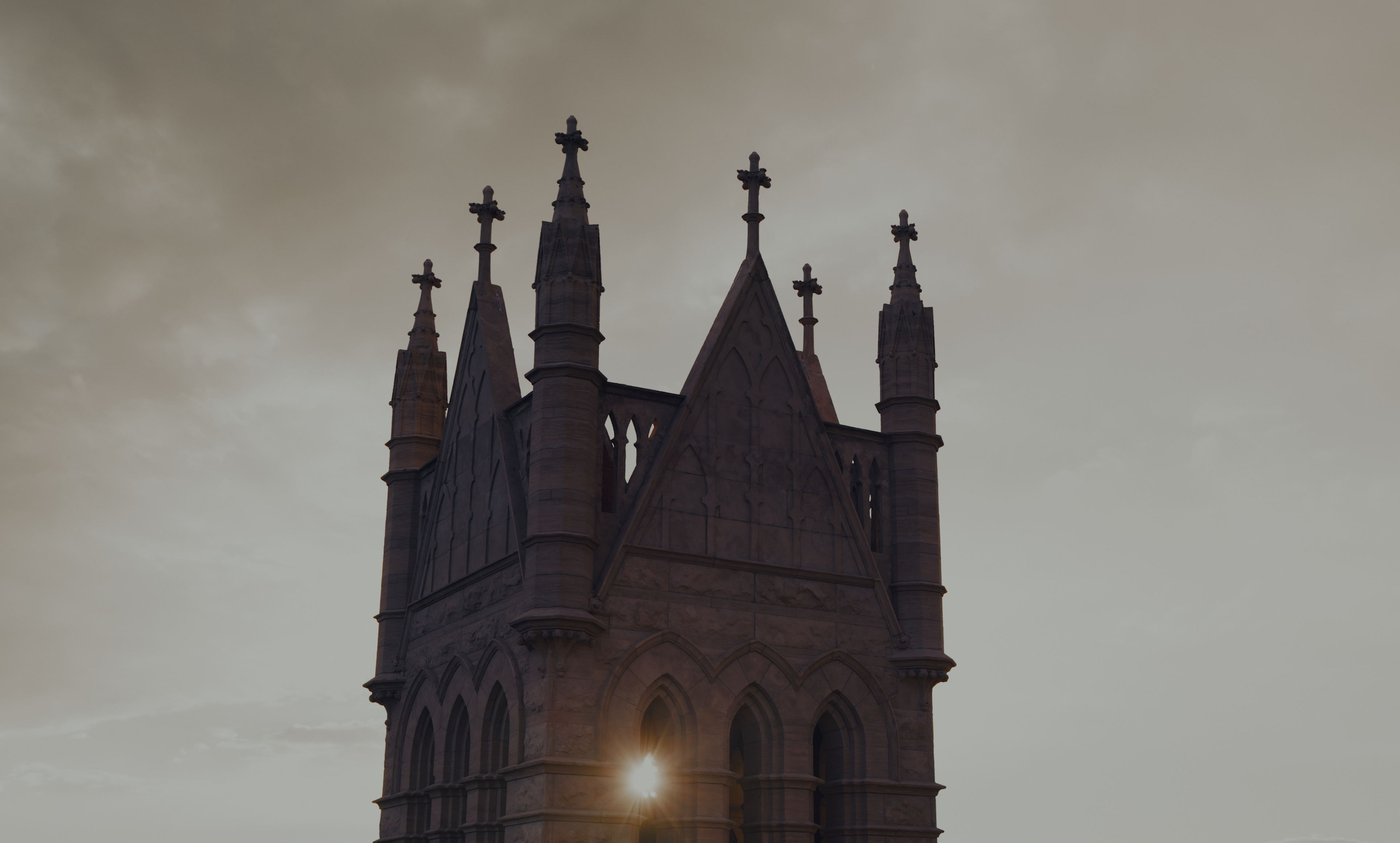 The gothic stone tower of a church against an overcast sky, sunlight flaring at its base