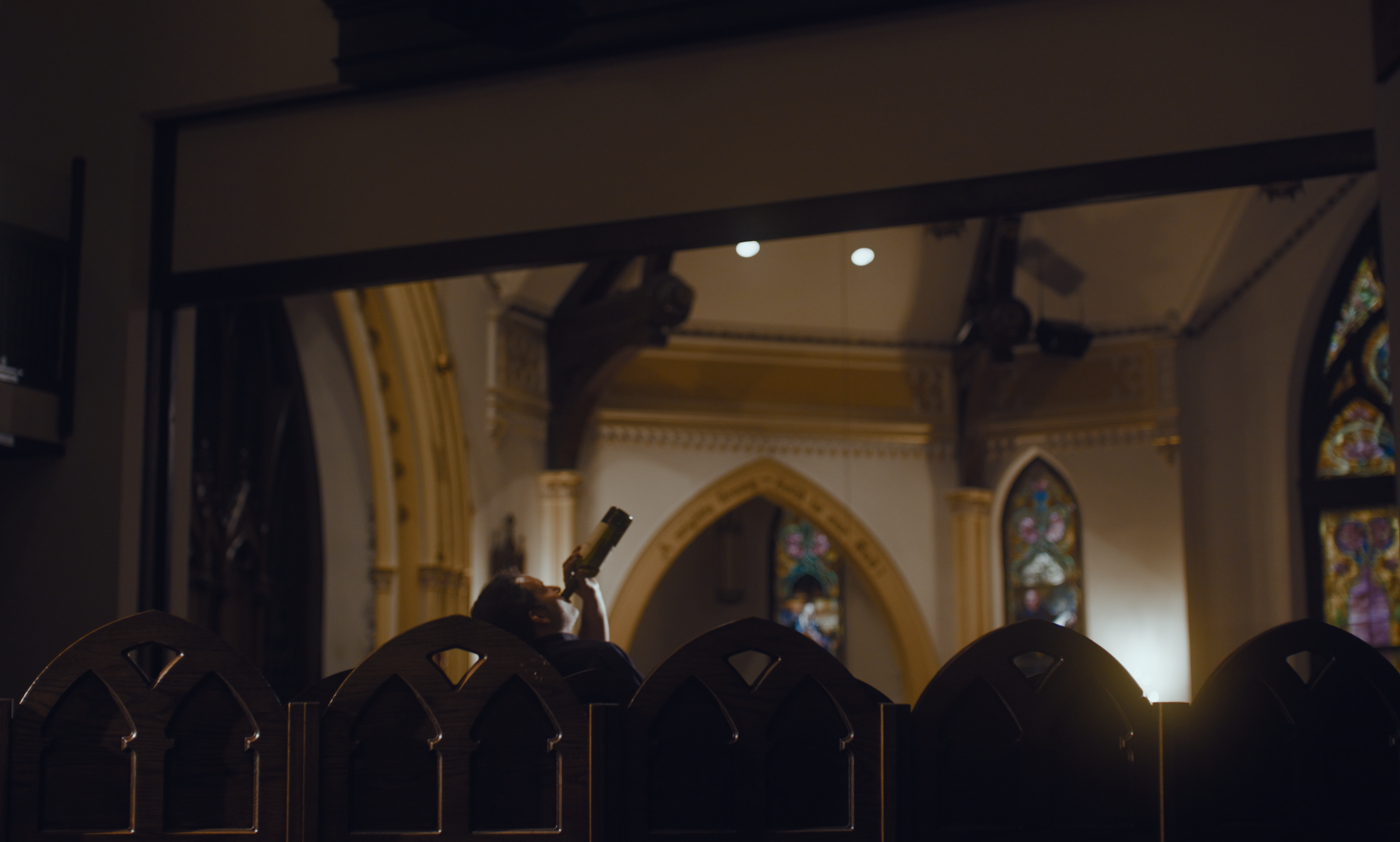 A man silhouetted behind the pews of a dim church, drinking from a bottle